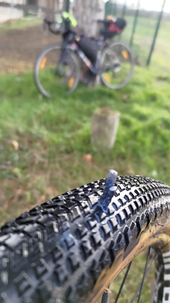 Gros plan sur un pneu de vélo dans lequel est enfoncée une mèche anti crevaison. À l'arrière plan avec un effet flou un autre vélo type course est posé sur un arbre