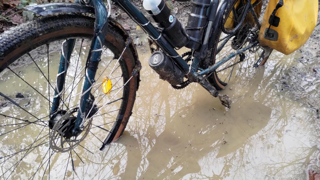 Vélo type gravel dans une flaque d'eau boueuse sur un chemin de forêt. L'eau de la flaque recouvre largement le bas des roues et la pédale pourrait être complètement sous l'eau en position basse.