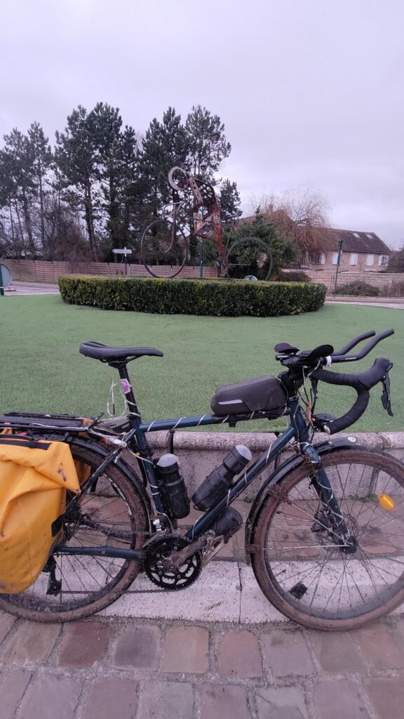 Vélo un peu boueux photographié sur un rond point devant une statue métallique représentant un cycliste et son vélo stylisé. C'est en hommage à Raymond Poulidor