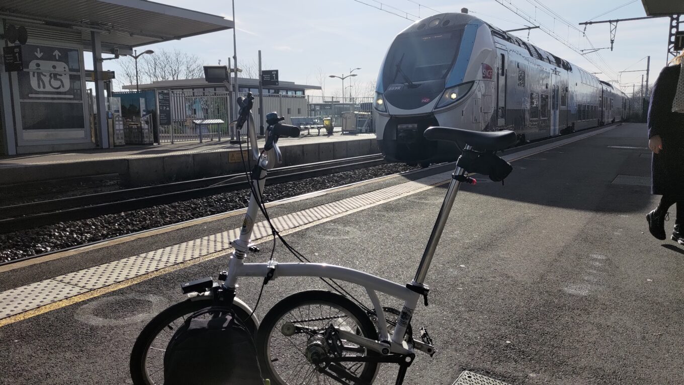 Vélo pliant à moitié plié sur un quai de gare, un train de banlieue arrive sur le quai