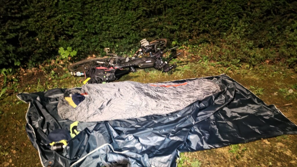 De nuit campement de fortune type bivouac, un tarp au sol, dedans bivi matelas duvet, vélo couché à côté du tarp