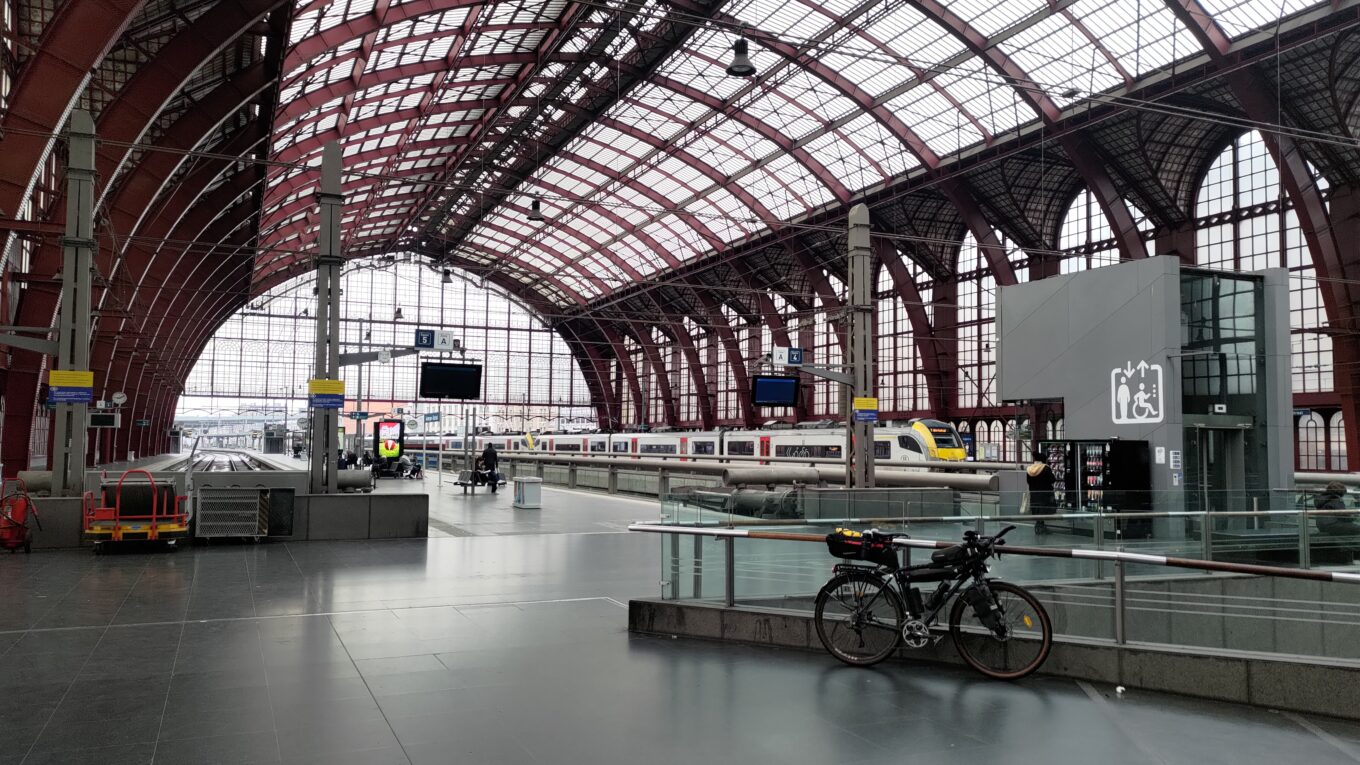 Vue de l'intérieur de la gare de Antwerpen, un vélo est posé sur un garde corps en verre, on voit la très grande verrière et les quais du premier étage de la gare, un train est visible sur l'un d'eux en arrière plan