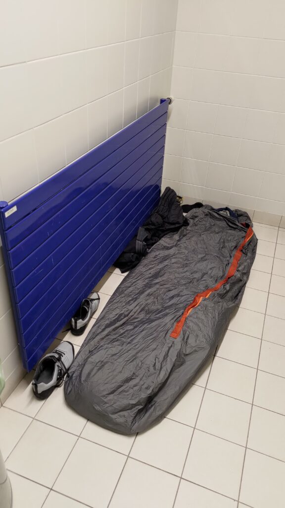 Dans un local spartiate, carrelage au sol et au mur (vestiaire dans un stade), un gros radiateur bleu au mur. Au sol de quoi passer la nuit, un bivy est visible et quelques affaires rangées sous le radiateur