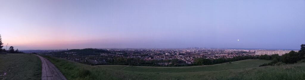 Vue panoramique depuis les hauteurs ez Argenteuil, levé de soleil sur Paris au petit matin. La vue ba de la défense sur la droite, la tour Eiffel au milieu et se perd ensuite dans l'est parisien. Un petit point dans le ciel montre la pleine lune. Le ciel varie di rose ai bleu