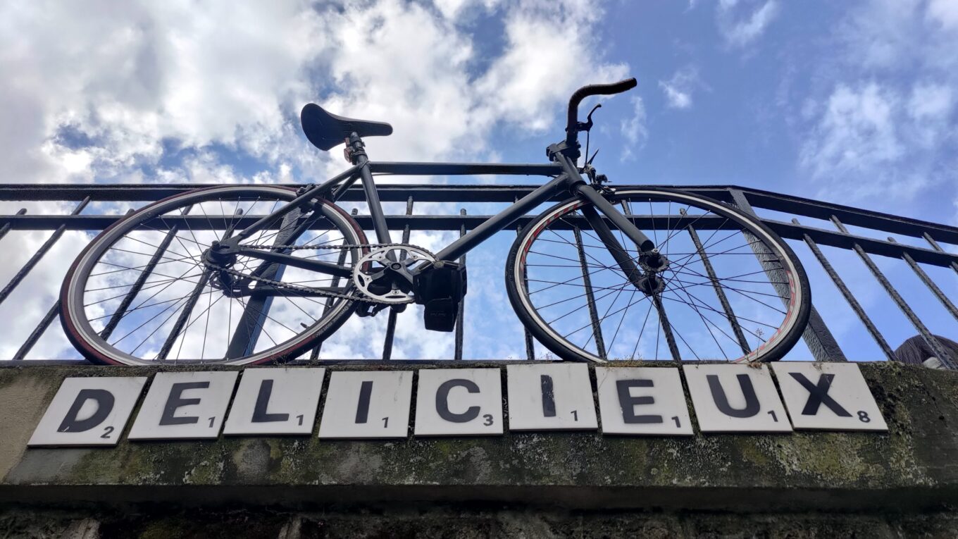 Vélo vu en contre plongée d'un vélo posé sur un parapet, dessous des lettres en céramique écrivant Délicieux
