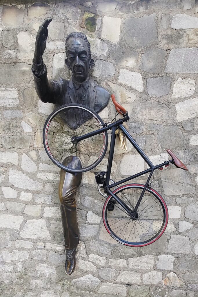 Statue du Passe Muraille à Paris (en bronze, plusieurs partie du corps sont incrustées dans le mur), un vélo suspendu sur sa main gauche