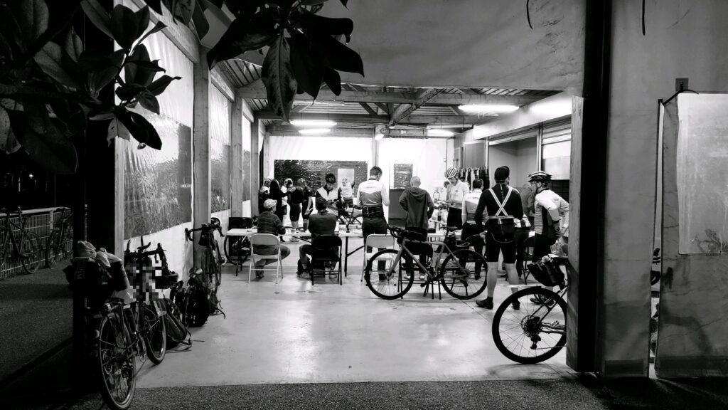 Sous un grand auvent, quelques tables, plein de vélo et de cyclistes. Photo en 'noir et blanc