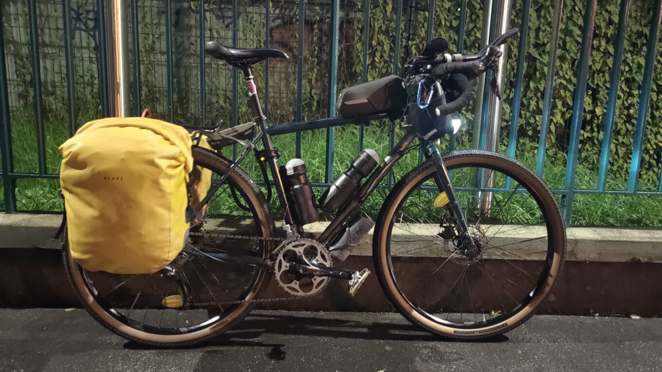 De nuit, un vélo type gravel avec des sacoches jaunes posées contre une barrière métallique