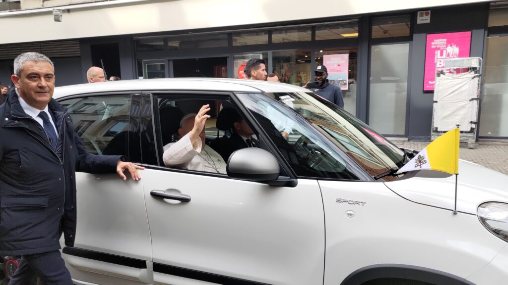 Le pape François saluant passant dans une rue en saluant depuis une petite voiture blanche. Le drapeau du Vatican est fixé sur la voiture, le visage du pape est masqué par sa main