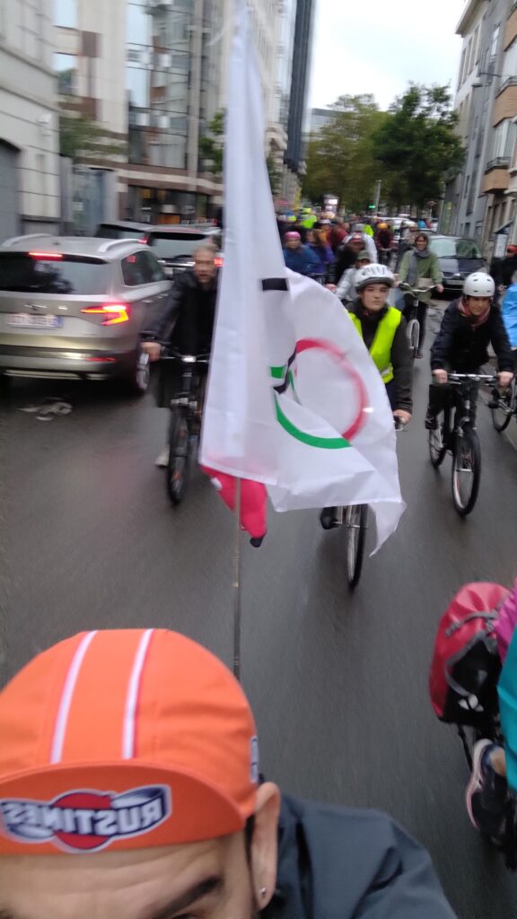 Selfie d'un cycliste dont on ne voit que le haut de la tête avec une casque orange, sur son vélo un drapeau olympique au centre de l'image, beaucoup de cyclistes derrière lui
