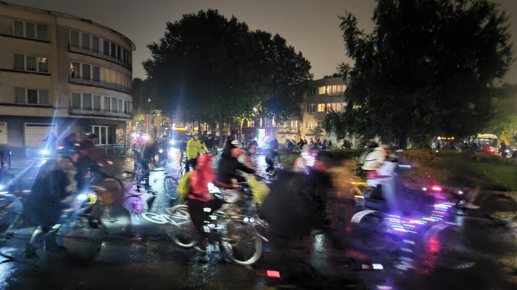 de nuit, de très nombreux cyclistes tournent autour d'une place. La photo est floue et l'on distingue les lumières qui brillent partout. Il pleut.