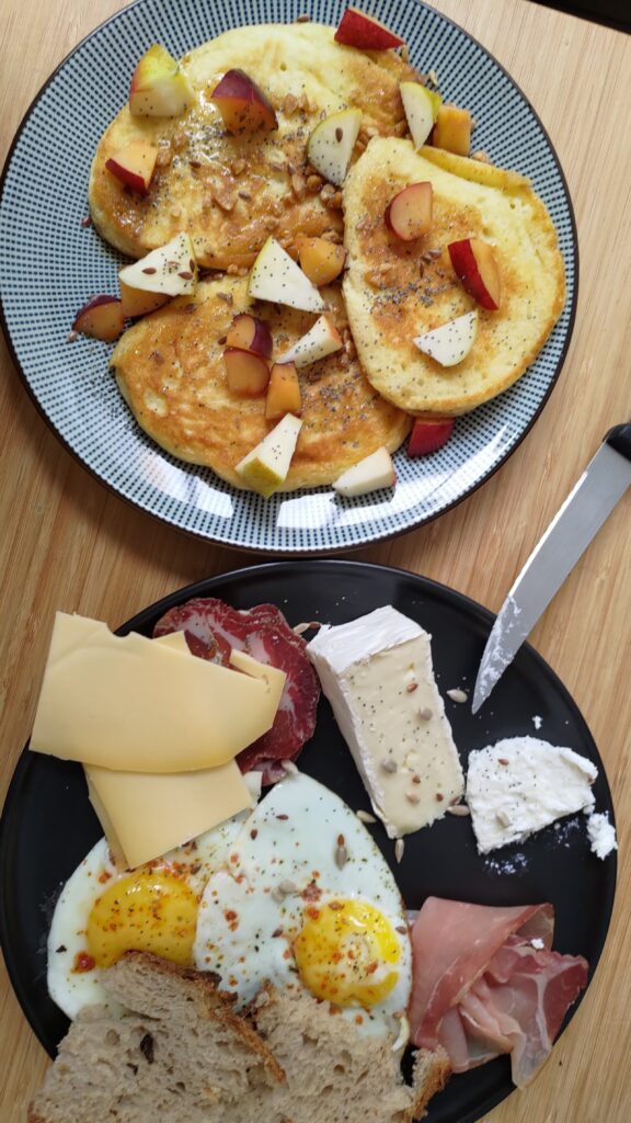 Brunch sur deux assiettes, l'une salée (pain œuf charcuterie fromage), l'autre sucrée (pancakes et morceaux de pommes)