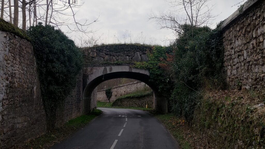 Une route et de part de d'autres de hauts murs de pierre d'une grand propriété. Un pont passe au-dessus de la route permettant de passer d'un côté à l'autre de la propriété