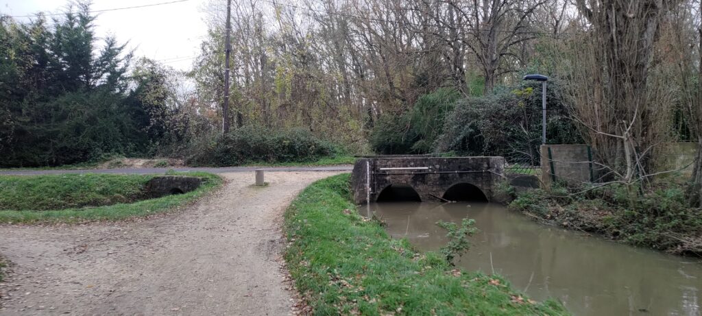 chemin de terre longeant une petite rivière qui passe sous un pont en pierres