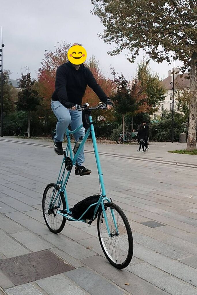 Une personne sur un tall bike cargo bleu, le visage est masqué par un smiley