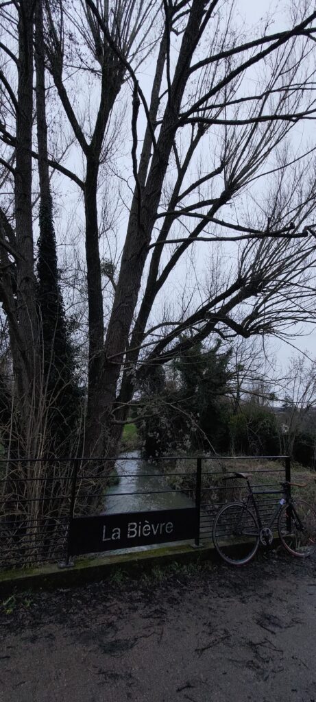 Petit pont avec une barrière métallique ornée d'une plaque avec écrit en creux La Bièvre. Derrière ine rivière et des arbres lz long. Sur la barrière est appuyé un vélo de tyoe fixie