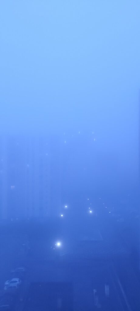 Vue depuis un immeuble en hauteur vers d'autres immeubles. L'image est toute bleue et on ne voit pas grand chose à cause du brouillard, les immeubles au fond sont à peine visibles, quelques lumières font des tâches lumineuses, on distingue des voitures garées