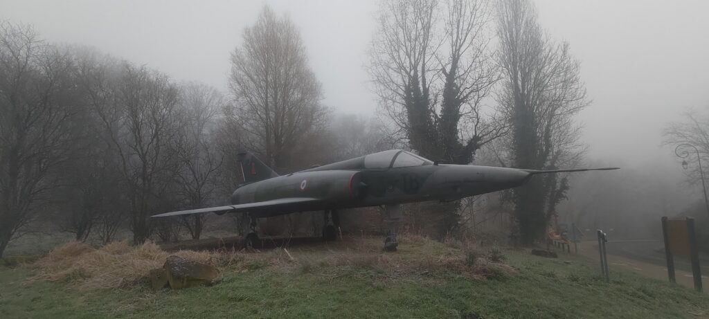 Un avion de chasse Mirage IIIR exposé en bord de route, dans le brouillard et ciel très gris. Il est couvert de givre