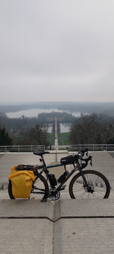 Un vélo type gravel avec des sacoches jaunes posé perpendiculairement à un paysage artificiel très rectiligne descendant une colline par des escaliers et prolongé au-dessus d'un plan d'eau par un pont suspendu rouge que l'on voir assez petit en contrebas mais qui paraît grand