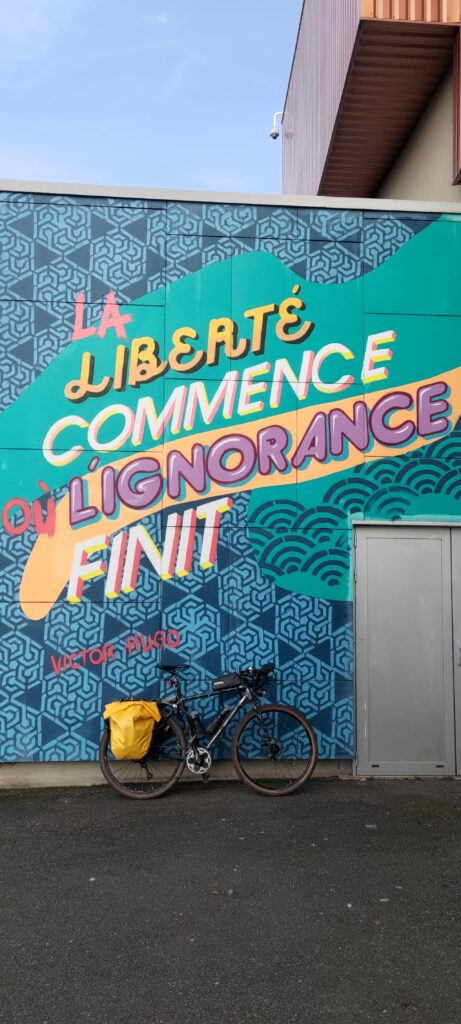Un vélo type gravel avec des sacoches jaunes posés devant un mur de graffitis faits sur un gymnase citant "La liberté commence où l'ignorance finit" signé Victor Hugo