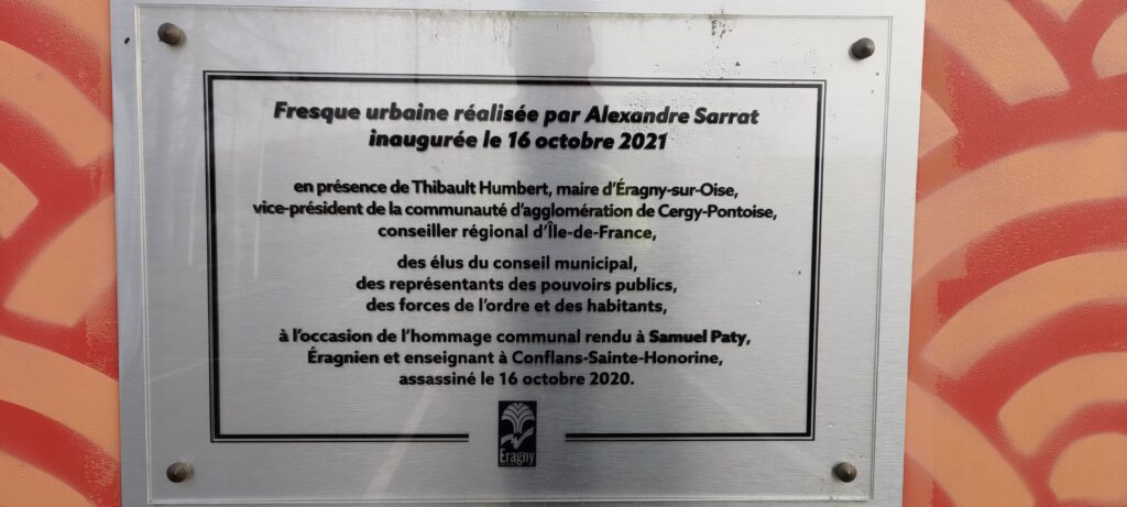 Plaque explicative des peintures murales d'un gymnase scolaire. Fresque urbaine réalisée par Alexandre Sarrat inaugurée le 16 octobre 2021 en présence de Thibault Humbert, maire d'Éragny-sur-Oise, vice-président de la communauté d'agglomération de Cergy-Pontoise, conseiller régional d'Île-de-France, des élus du conseil municipal, des représentants des pouvoirs publics, des forces de l'ordre et des habitants, à l'occasion de l'hommage communal rendu à Samuel Paty, Éragnien et enseignant à Conflans-Saine-Honorine, assassiné le 16 oectobre 2020.