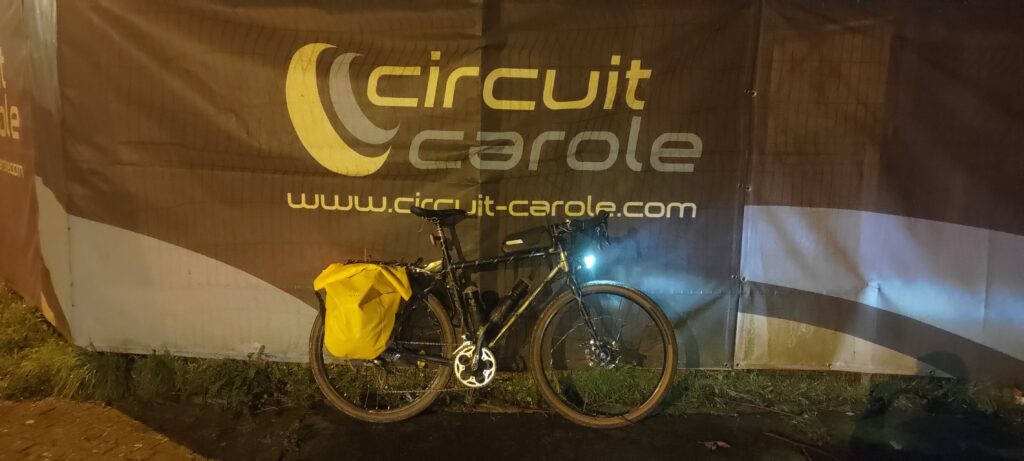 De nuit, un vélo type gravel avec des sacoches jaunes, lumières allumées, posé sur un grillage recouvert d'une bâche décorée du logo Circuit Carole