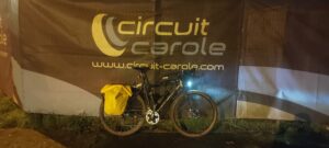De nuit, un vélo type gravel avec des sacoches jaunes, lumières allumées, posé sur un grillage recouvert d'une bâche décorée du logo Circuit Carole