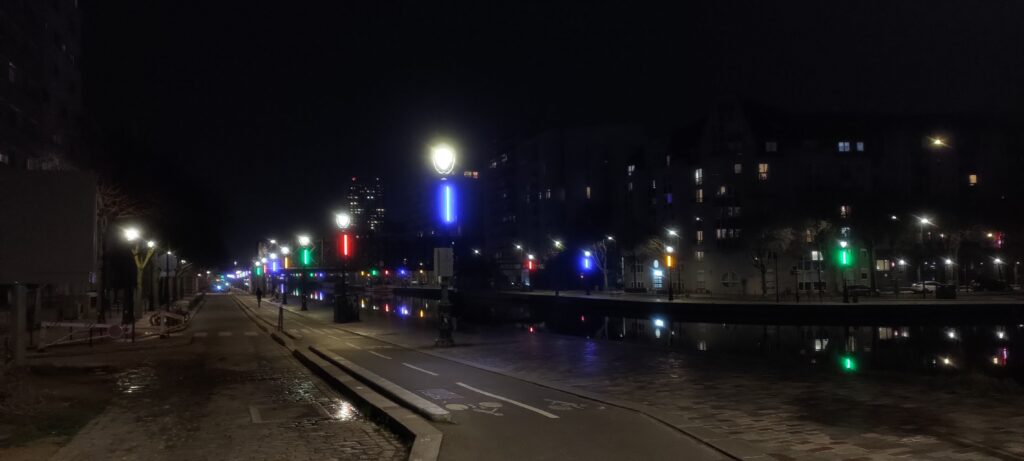 De nuit, kes bords d'un canal dont les deux rives sont éclairées de lampadaires décorés de lumières vives bleue/rouge/verte/jaune. une piste cyclable longe le canal, une petite rue longe la piste.