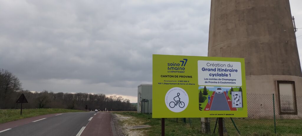 Un panneau d'information en bord de route indiquant la Création du Grand itinéraire cyclable 1 par le département de la Seine et Marne, la route est une chaussidou neuve avec la partie voiture en noir et la partie cycle en rouge