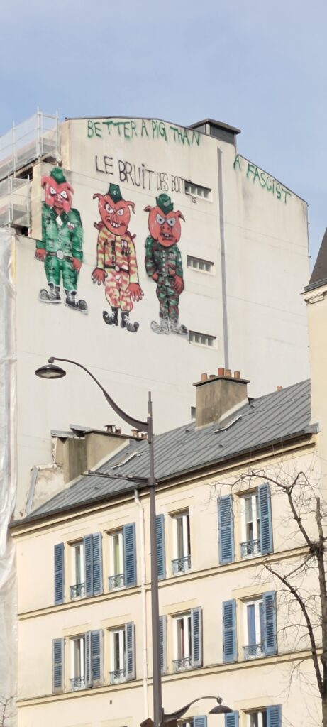 Sur un mur pignon d'immeuble, sur les 3 derniers étages, un texte le long du toit "BETTER A PIG THAN A FASCIST", dessous "LE BRUIT DES BOTTES" et dessous trois cochon humanoïdes en tenue militaires différentes et l'air à la fois agressif et ahurit.