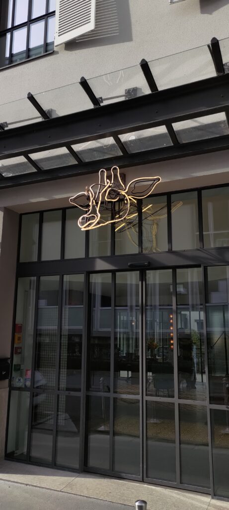 Entrée en verre d'un bâtiment surmontée d'un néon lumineux représentant une tête de girafe stylisée