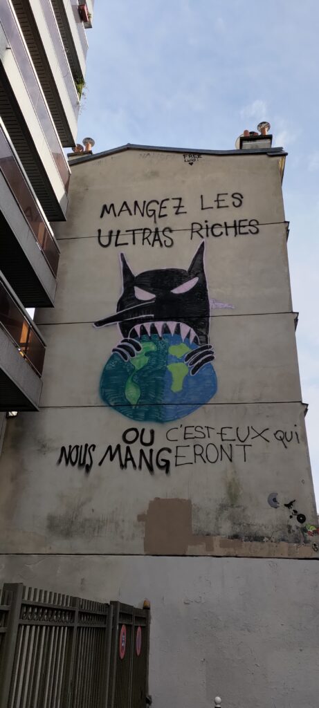 Sur un pignon de mur d'un immeuble de 6 étages environ, un monstre tout noir avec des grandes dents pointues mets dans sans bouche la planète Terre. Texte "Mangez les ultras riches ou c'est eux qui nous mangeront"
