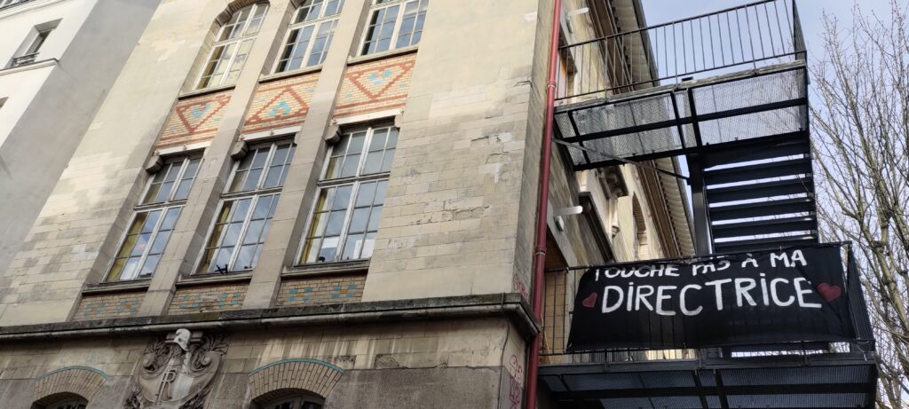 Sur une école, un grand escalier métallique le long de la façade et une bâche "Touche pas à ma directrice" avec des cœurs de chaque côté du mot "directrice"