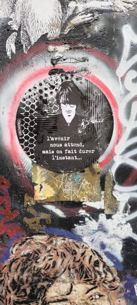 Gros plan sur un mur graphé et avec un collage d'un jeune homme à lunette, cheveux mi-longs qui lui recouvre le visage qu'il penche vers le bas et lunette rondes. "L'avenir nous attend, mais on fait durer l'instant…"