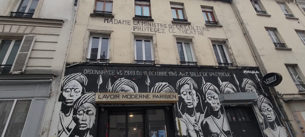 Façade d'un immeuble parisien, entre le 2ème et le troisième étage sur 4 fenêtres de large texte "MADAME LA MINISTRE DE LA CULTURE PROTÉGEZ LE THÉÂTRE". Au rez de chaussée "Ordonance 45.2339 du 13 octobre 1948 Art2 Salle de Spectacle" et portraits géants au niveau du torse de 9 soldats de type Massai avec turban et lance. Entrée "LAVOIR MODERNE PARISIEN"