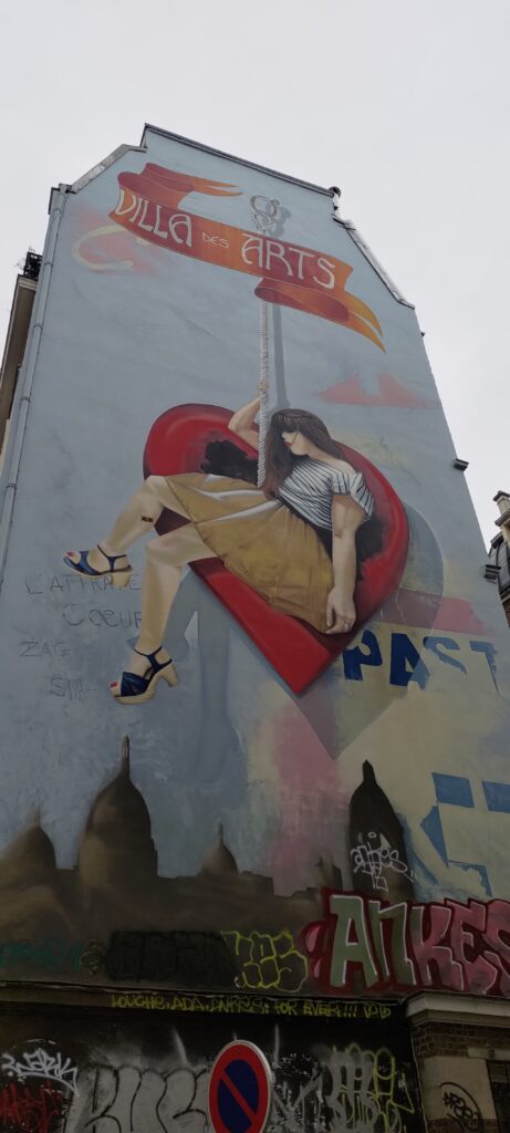 Sur un pignon de mur d'un immeuble d'environ 6 ou 7 étages, une femme en jupe marron et marinière blanche et bleue est accrochée à une corde qui maintient également un cœur géant sur lequel elle est assise. Texte en haut sur un ruban "Villa des Arts" (police art déco)