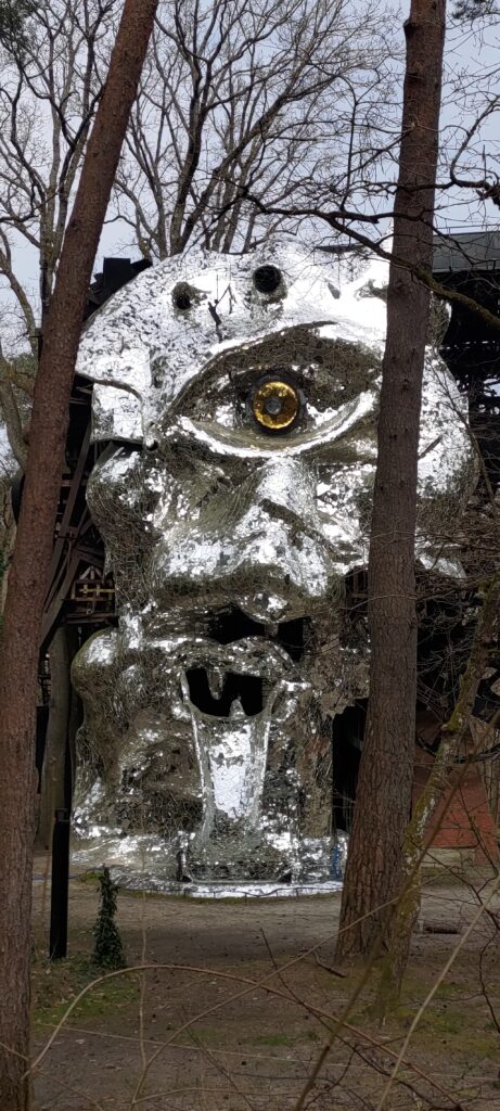 Sculpture métallique ou en miroir de plusieurs mètre de haut représentant un visage torturé avec un unique œil jaune. Le Cyclope