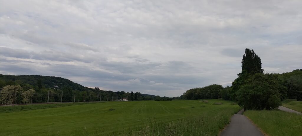 Paysage de campagne, une voie de chemin de chemin de fer à gauche, un champs au centre qui occupe la majorité de l'image, à droite une piste cyclable (et on devine une route encore plus à droite)