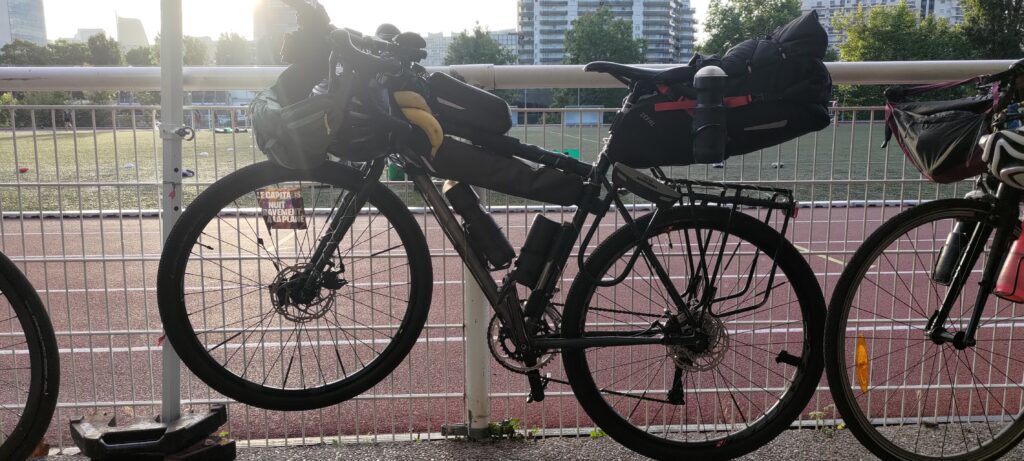 Un vélo type gravel chargé en bagages (et avec deux bananes) suspendu par le guidon à la barrière d'un stade