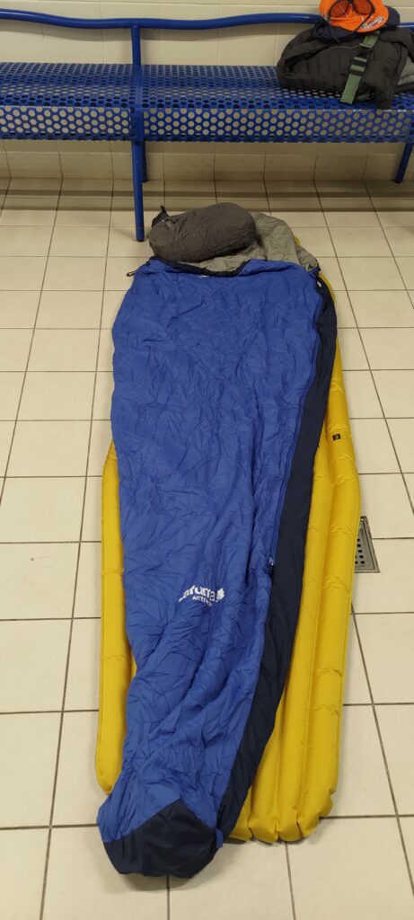 un matelas et un sac de couchage posés au sol sur du carrelage dans un vestiaire