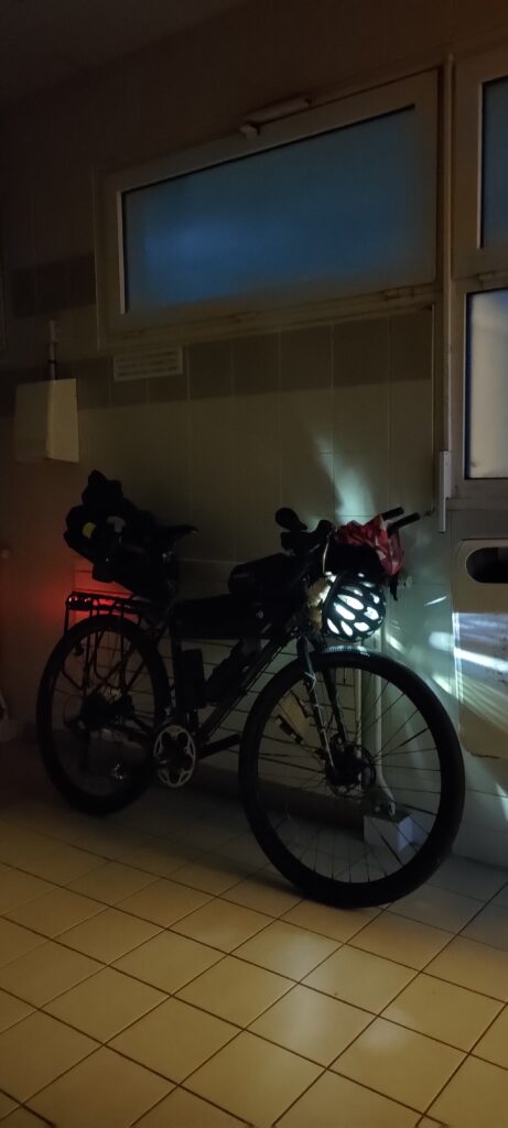 Un vélo type gravel, de nuit dans le local faiblement éclairé. Les lumières du vélo sont allumées La lumière avant filtre à travers le casque cycliste posé devant le phare.