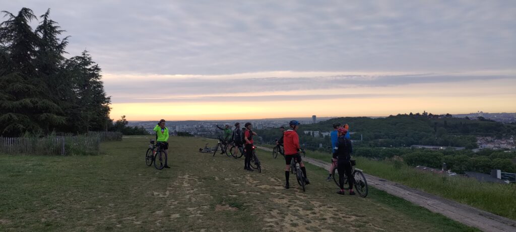Groupe de cyclistes au levé du soleil sur les hauteurs d'une butte, on distingue Paris au loin