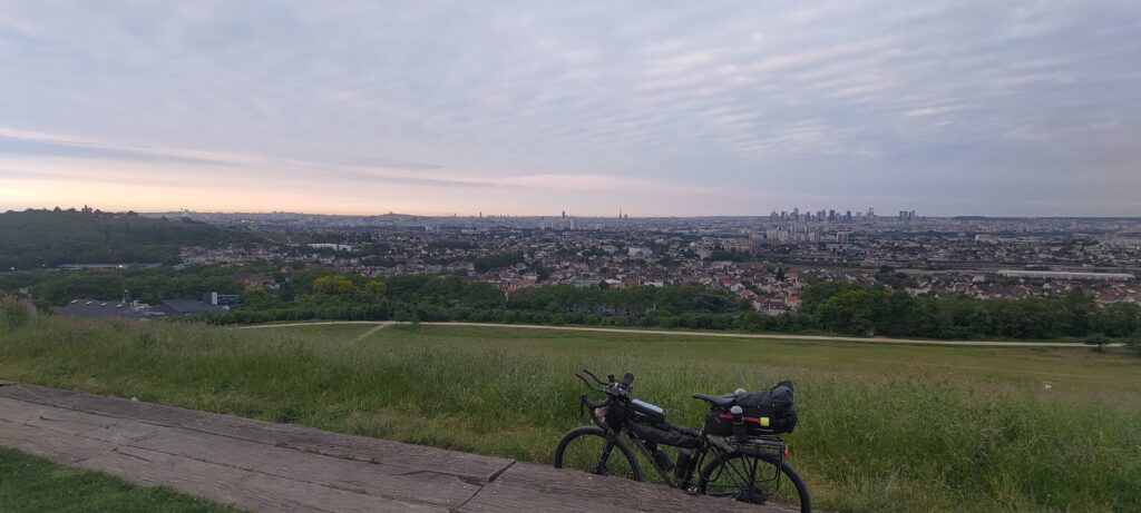 Un vélo type gravel en haut d'une butte, à l'horizon Paris où l'on distingue en plein centre la tour Eiffel, sur la gauche la butte Montmartre et le Sacré Cœur, et sur la droite les tours de la Défense