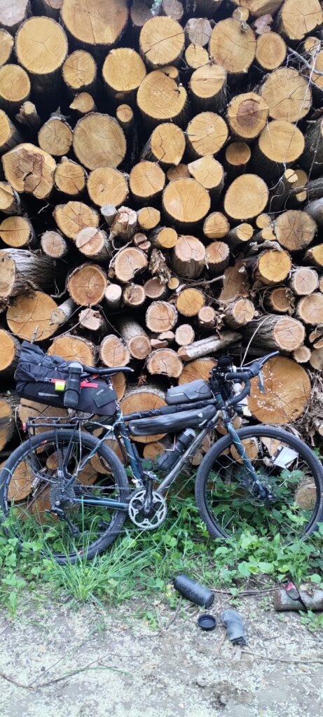 Un vélo type gravel appuyé sur une très haute pile de bois dont on ne distingue pas le haut. Au pied du vélo des affaires dont une trousse à outils