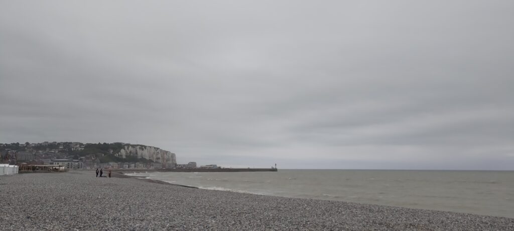 Plage de galets gris, mer grise, falaise au loin et ciel gris