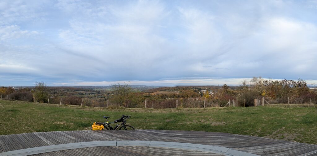 Vue panoramique depuis le haut de la colline d'Élancourt, la table d'orientation qui est une très grande terrasse en bois avec un cercle métallique au sol indiquant les points d'intérêts est au premier plan, un vélo est appuyé dessus