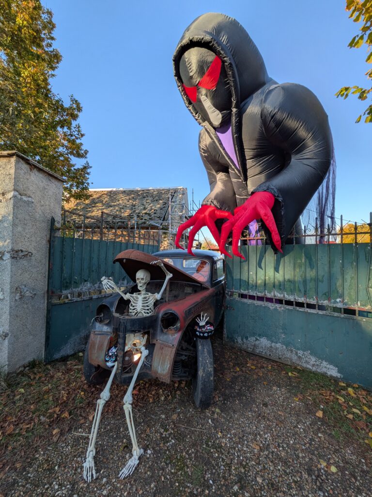 Maison décorée pour Halloween, une vieille voiture toute rouillée sort d'une maison dont le portail est fermée sur la voiture, sortant du capot avant, un squelette dont les jambes sont à l'emplacement du radiateur. Sur la voiture plusieurs bac à bonbons. Au-dessus du portail, une structure gonflée d'un personnage effrayant, grandes mains griffues rouges, yeux rouges, long manteau noir. Il est 2 fois plus haut que le portail