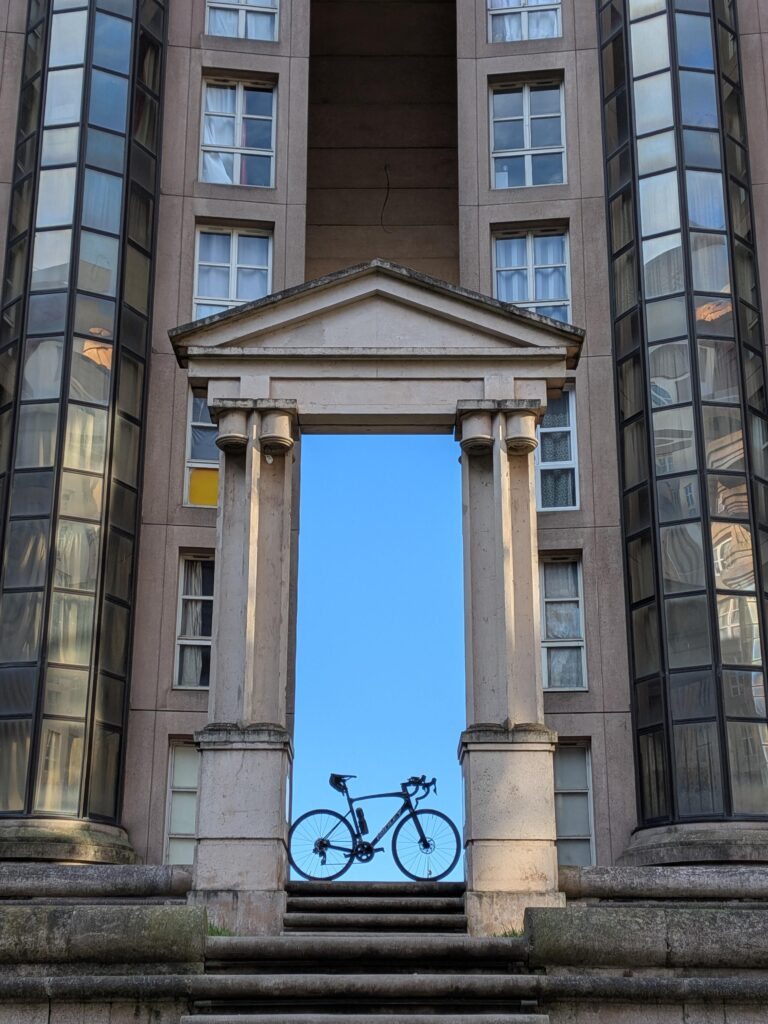 Au cœur d'un immeuble d'habitation de style brutaliste, béton et verre, une porte d'aspect antique laisse passer un ciel bleu. Un vélo de course est posé sous cette porte et son ombre se découpe sur le ciel, en vue serrée, la porte occupe presque la totalité de l'image