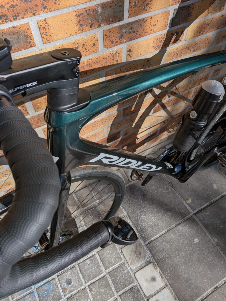 vue en plongé sur un vélo de course vert foncé et noir, on distingue une partie du cintre et l'avant du cadre. il est posé sur un mur en briques