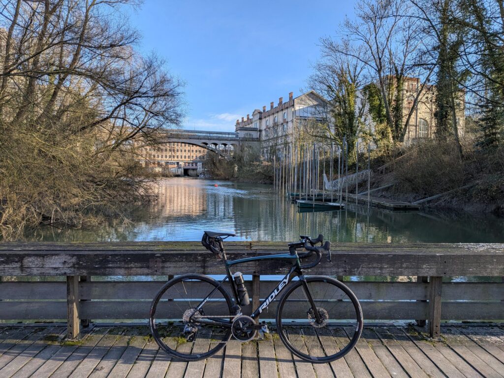 un vélo de course sur un pont en bois au dessus d'une rivière, en arrière plan un bâtiment industrielle et un grand pont métallique qui traverse la même rivière. Des arbres sans feuille bordent le court d'eau de part et d'autres